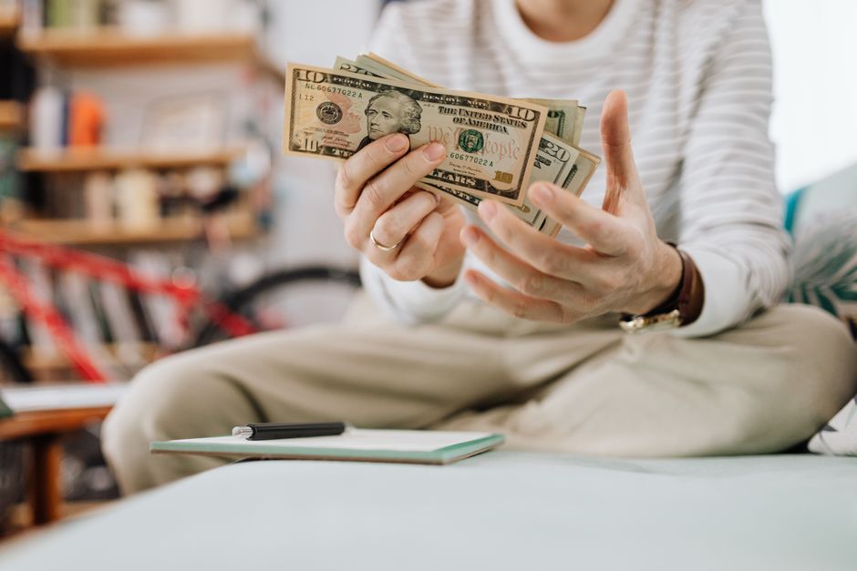 A person sitting cross-legged holding a small fan of US dollar bills, a notebook and pen on the seat, a red bicycle blurred behind