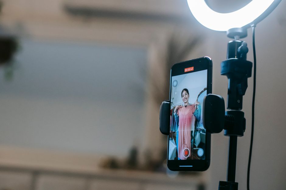 A smartphone clamped to a tripod beside a lit ring light, its screen showing a woman holding up a pink dress while recording, timer at 47 seconds, blurred room behind