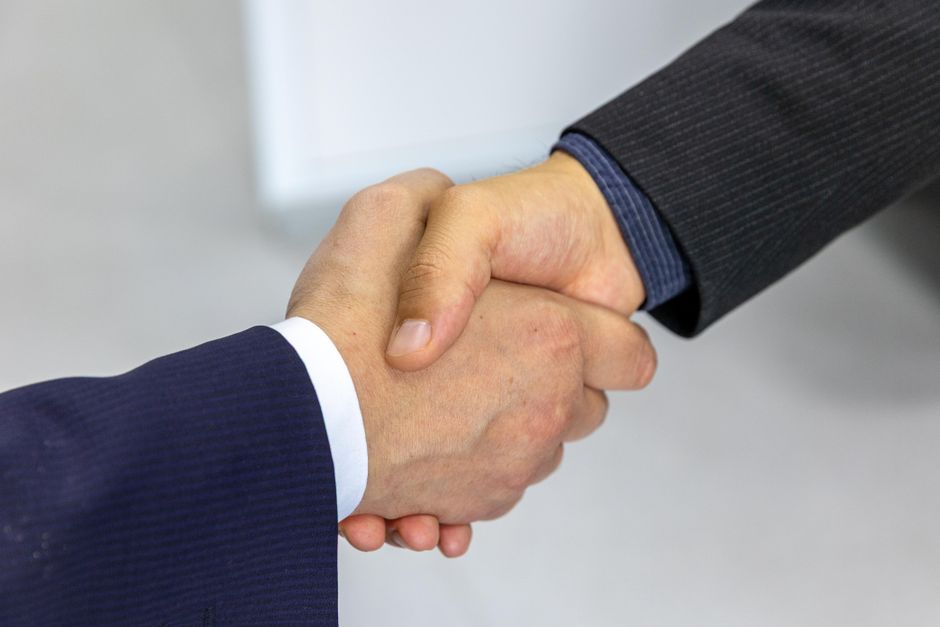 Two people in dark suits shaking hands against a plain light background, only forearms and hands visible