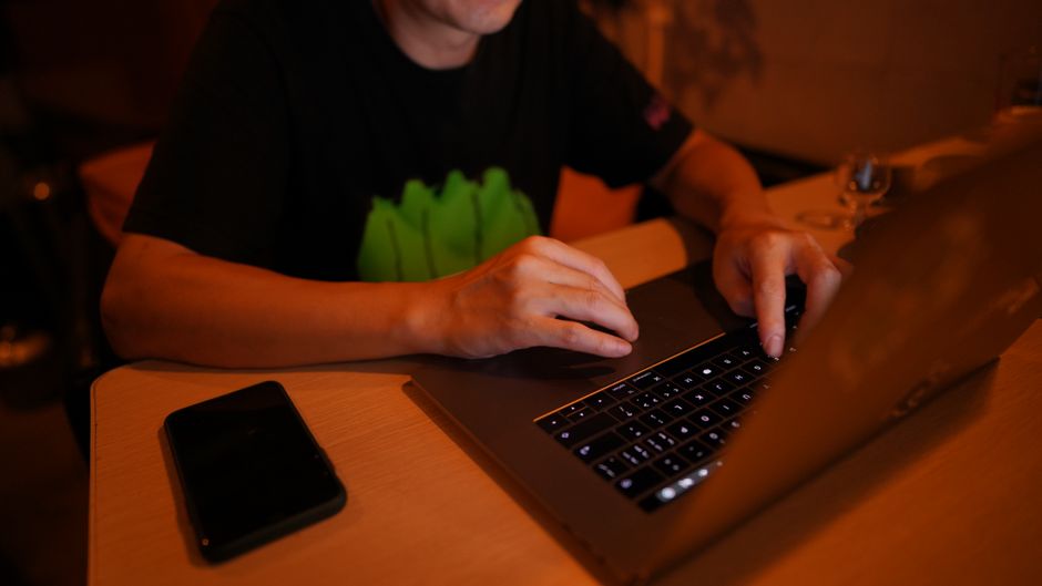 Close-up of a man's hands typing on a laptop keyboard in warm orange light, a phone face-down on the wooden desk beside him