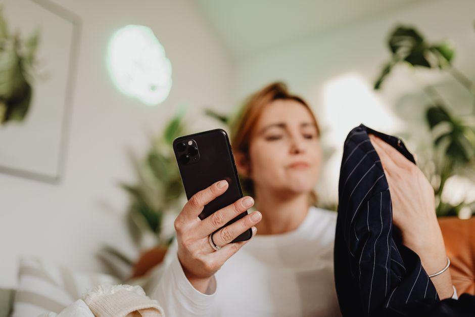 A woman lying on a sofa looking at her phone while holding up a navy pinstriped jacket, green plants behind her