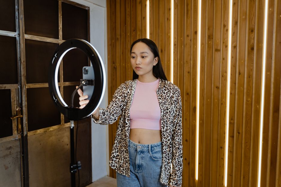 A smartphone clamped to a tripod beside a lit ring light, its screen showing a woman holding up a pink dress while recording, timer at 47 seconds, blurred room behind